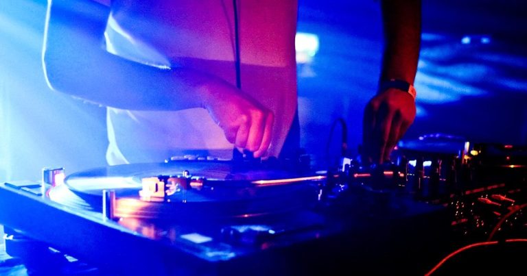 A DJ is mixing music on a turntable in a dimly lit environment with blue and pink lights. The image focuses on their hands adjusting records and controls, with various knobs and equipment visible.