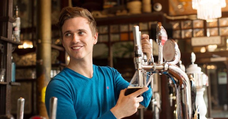 A person in a blue sweater is smiling while pouring a drink from a tap at a bar in a pub. The background shows bar shelves and decorations.