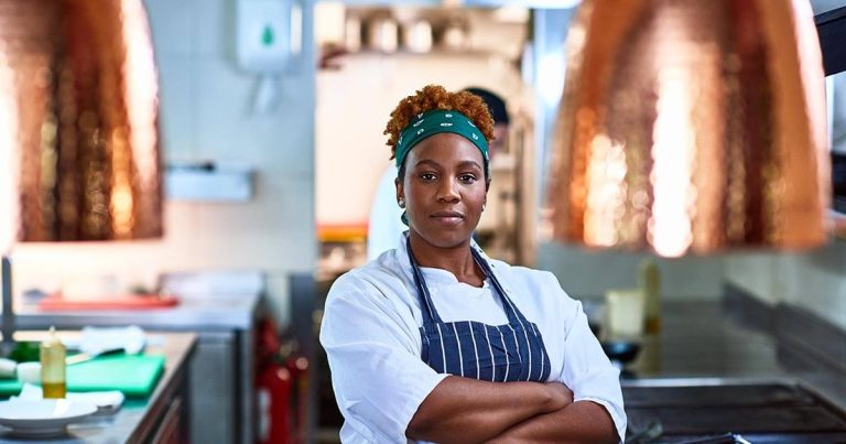 A chef wearing a white coat and striped apron stands confidently in a commercial kitchen. Her hair is tied back with a green headscarf. Copper pendant lights hang above, and kitchen equipment is visible in the background.