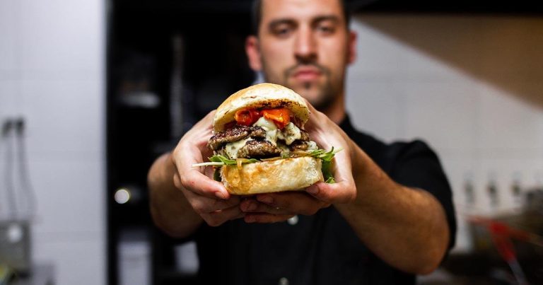 Man holding a large sandwich in a kitchen with grilled meat, cheese, and vegetables between a bun, background blurred.