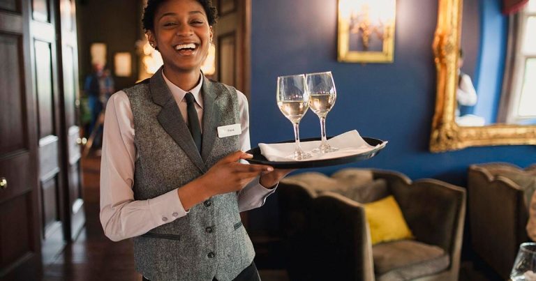 A person wearing a gray vest and white shirt holds a tray with two glasses of white wine. They are standing in a warmly lit room with blue walls and vintage decor, including a large gold-framed picture. The person is smiling.