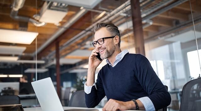 Man talking on smartphone at a desk in a modern office while facing his open laptop.
