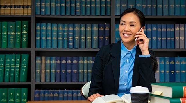 A woman in a blue shirt and black blazer is talking on a smartphone. She is sitting at a desk with an open book, a coffee cup, and more books in the foreground. Behind her are shelves filled with blue and green books.