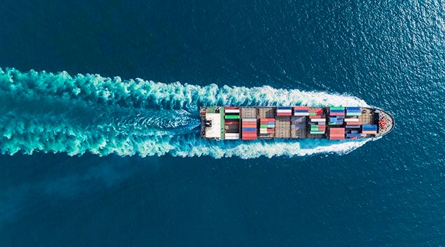 Aerial view of a large cargo ship moving through deep blue ocean waters, leaving a trail of waves behind. The ship is loaded with multiple colorful shipping containers arranged in neat rows.