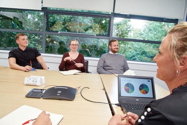Group of four people in a business meeting. A woman has a laptop open, and it is showing a pie chart slide.