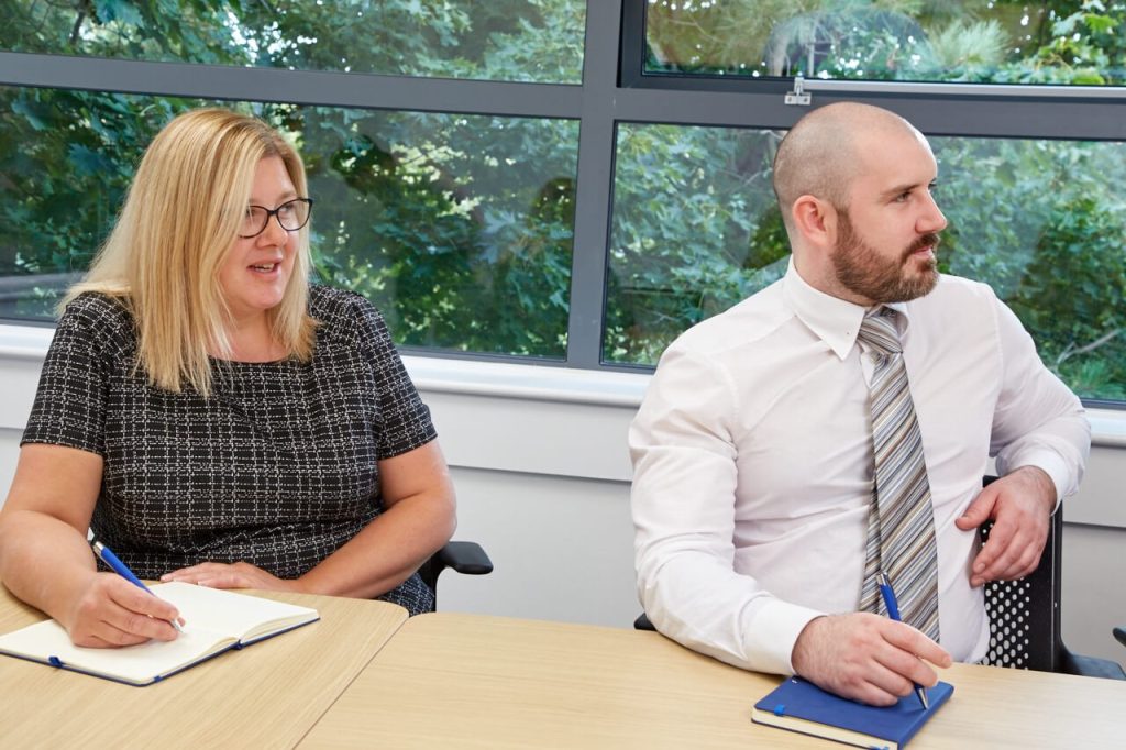 Two people taking notes at a conference table, with a window showing trees outside.