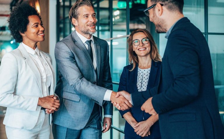 Four people in business attire shaking hands in hallway of modern office.
