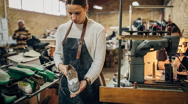 Person inspecting a shoe in a workshop with shoemaking tools and machinery in the background.