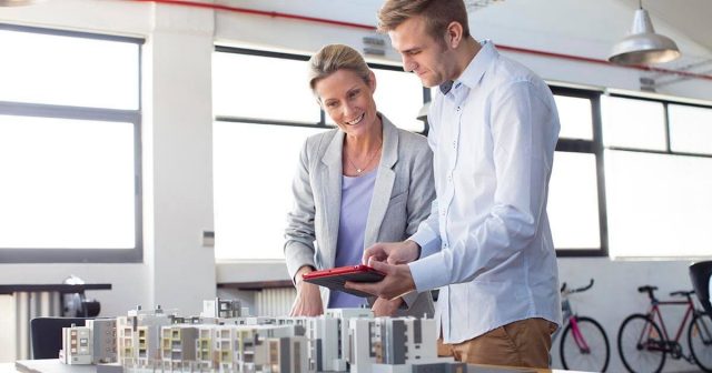 Two people examine architectural models on a table, sunlight streaming through large windows.