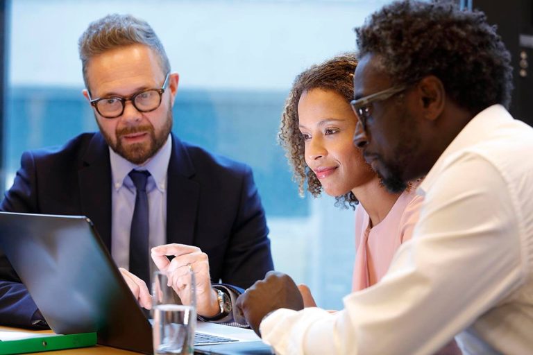 Three professionals sitting at a meeting table, discussing, with a glass of water on the table and a laptop open.