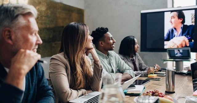 Group of four people in a meeting room with a video call on a large screen at the table.
