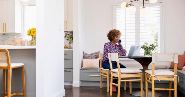 A business consultant sitting at a round table with a laptop open while talking on a smartphone while working from home.