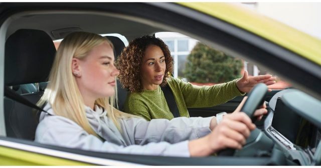 Two women are in a car; the woman in the driver's seat is holding the steering wheel while the other is providing instructions.