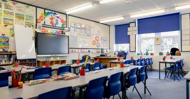 Classroom with desks, digital display, and educational posters, and supplies organized on desks and a bookshelf.