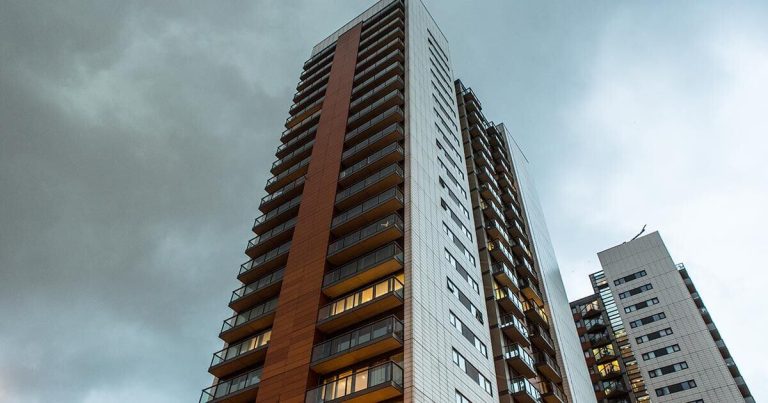 Tall residential buildings with multiple balconies under a cloudy sky. The structures have a modern design, featuring sections with different materials and colors. Some windows are illuminated, suggesting an evening setting.