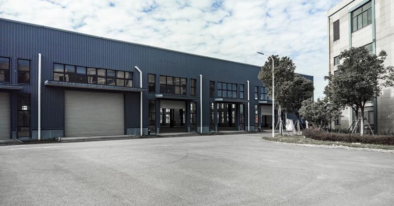 A large industrial building with blue metal siding and several wide roll-up doors. The area is empty, with a paved driveway in front. There are trees along the side and a modern office building on the right. The sky is cloudy.