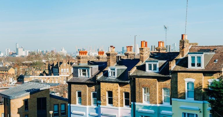 Row of brick townhouses with dormer windows, and a city skyline with cranes in the background.
