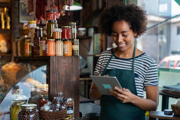 Smiling deli worker using a tablet, surrounded by shelves of jars.