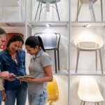 Three people in a furniture shop are standing by a shelving unit with chairs, looking at a tablet that one of them is holding.