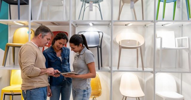 Three people in a furniture shop are standing by a shelving unit with chairs, looking at a tablet that one of them is holding.