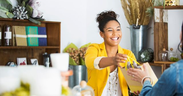 A smiling woman in a yellow blazer hands a shopping bag to a customer in the store, where shelves with decorative items and plants create a warm and inviting ambiance in this cozy shop .
