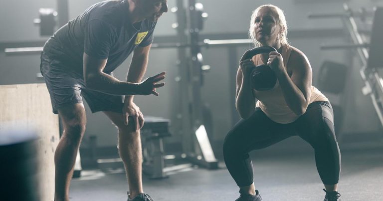 Woman performing a squat with kettlebell, coached by a man in a gym setting.