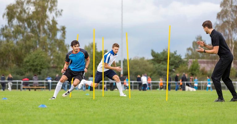 Two football players running through a training drill with yellow poles and a coach clapping.