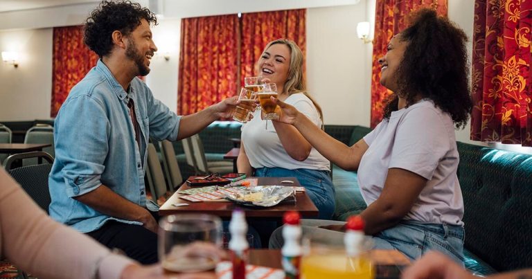 Three people toasting with beer glasses in a social club, with snacks on the table.