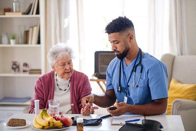 A healthcare professional in blue scrubs is checking the blood sugar of an elderly woman seated at a table adorned with fruits, bread, and a glass of water. In this well-lit room with shelves behind them, the peace of mind from care home insurance ensures their environment remains secure and supportive.