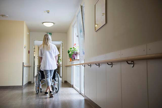 A healthcare worker pushes a person in a wheelchair down a well-lit hallway, where the cream-colored walls symbolize the comfort and safety ensured by care home insurance. Handrails line both sides, with a potted plant on a window ledge adding warmth to this secure environment.