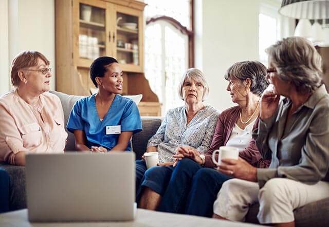A group of five older women sit on a couch having a conversation. One woman, in a blue nurse uniform and name tag, discusses nursing home insurance while the others listen intently. A wooden cabinet and window are in the background. A laptop is open on a table nearby.