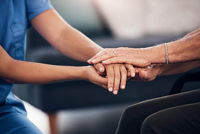 A healthcare worker in a blue uniform gently holds the hands of an elderly person, symbolizing the attentive care often assured by nursing home insurance, with the focus on their hands against a blurred background.