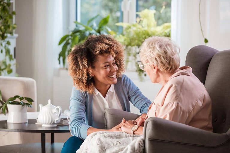 A young woman with curly hair is sitting on a chair, smiling and holding the hands of an elderly woman with glasses in an armchair. In this cozy, plant-filled room that feels like a haven, nearby sits a teapot and cups—a warm moment supported by reliable care home insurance.