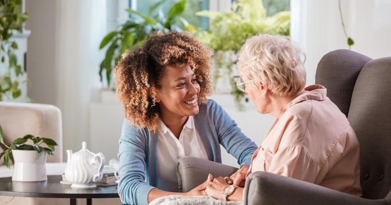 A younger woman with curly hair smiles while holding the hands of an older woman with short hair seated in a chair. They are indoors at a cozy care home, surrounded by plants, with a teapot on the table nearby—a scene that highlights the warmth and security supported by care home insurance.