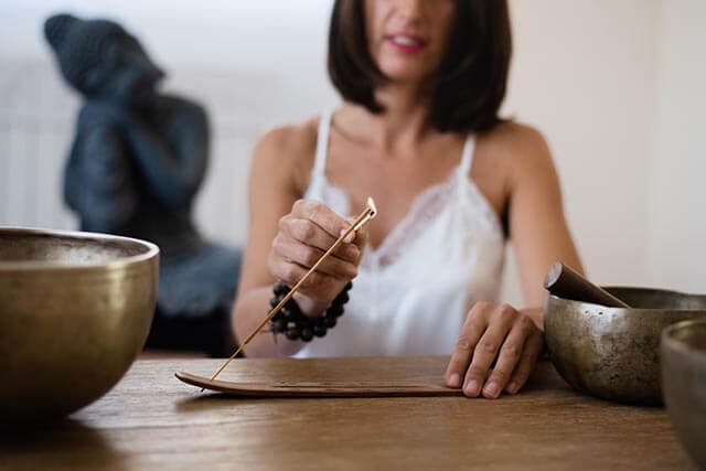 A woman in a white top is sitting at a wooden table, holding a lit incense stick over a holder. There are two brass bowls on the table. A statue is visible in the background.