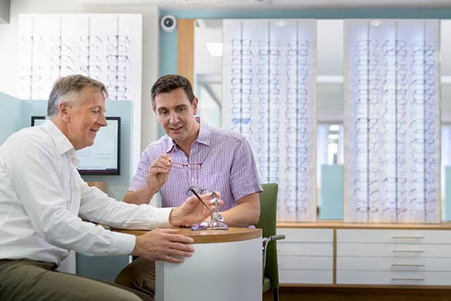 Two men sit at a small table in an opticians store. One man, in a white shirt, points at a tablet, while the other, in a purple shirt, holds a pair of glasses. Display shelves with rows of eyeglasses are visible in the background.