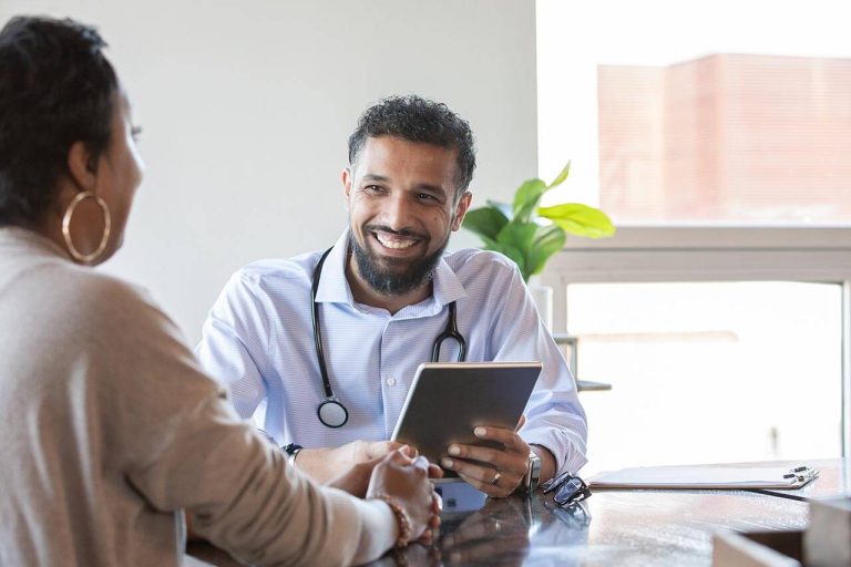 Doctor holding a tablet, smiling while consulting a woman in a medical office.