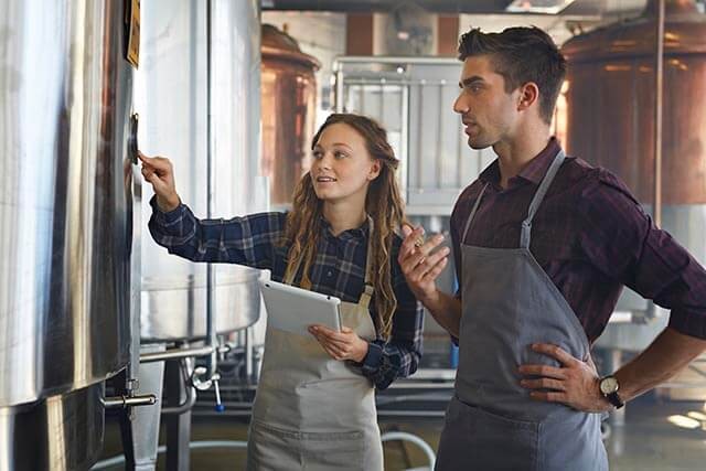 Two people in aprons stand in a brewery. One holds a tablet and points at a metal container, while the other stands with a hand on their hip. Industrial equipment is visible in the background.
