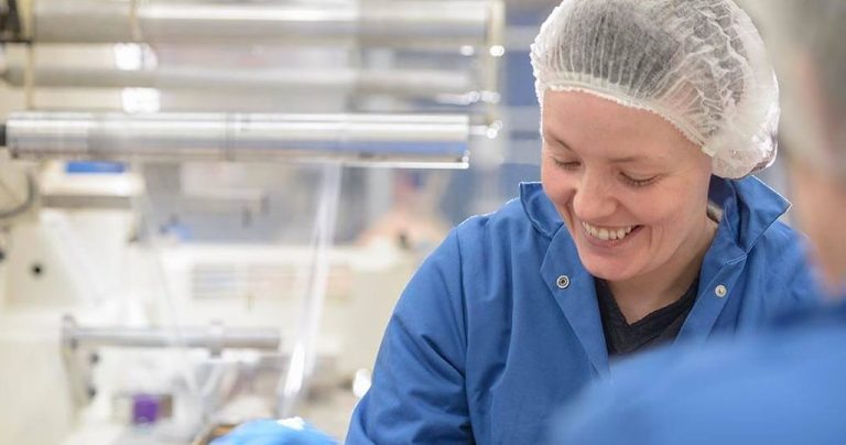 Worker smiling in a food manufacturing facility, wearing a lab coat and hairnet with equipment in the background.