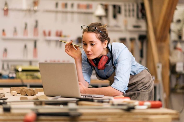 Person in a furniture workshop using a laptop, surrounded by tools and equipment.