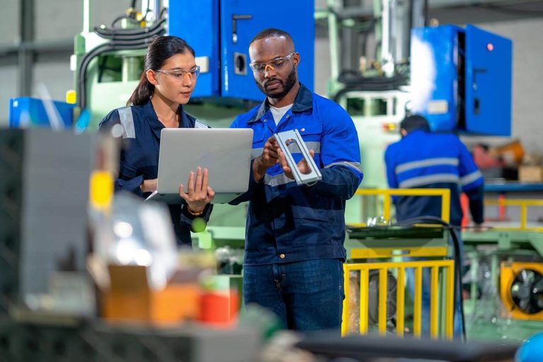Two factory workers in a manufacturing facility, examining machinery while one of them is holding a laptop.
