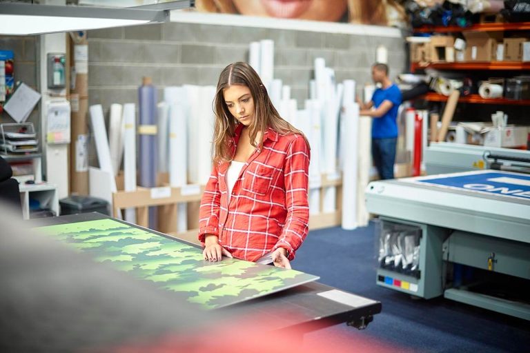 A woman works at a table with green and black patterned material. There is printing equipment in the background.