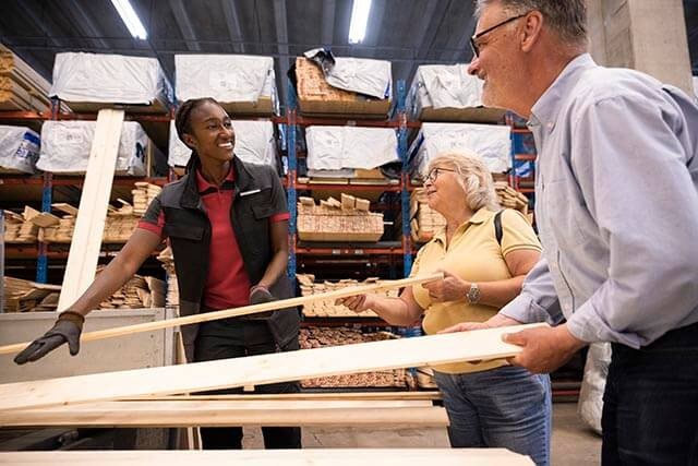 Timber merchant discussing with customers in a lumber warehouse with more stacked materials on the shelves in the background.