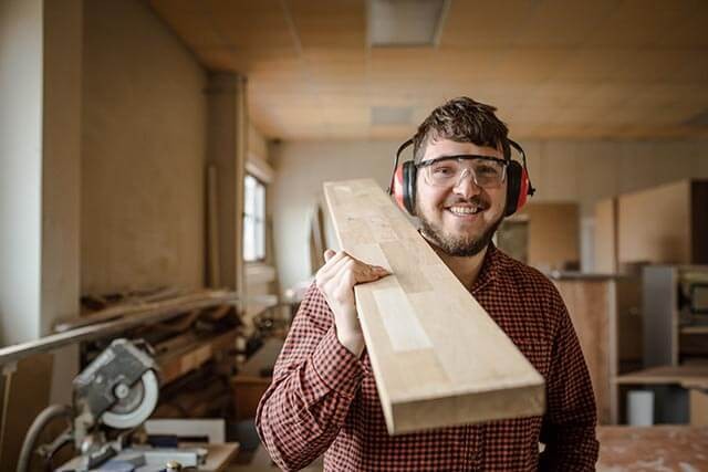 Person holding a wooden plank over his shoulder in a workshop, wearing ear protection and safety goggles.
