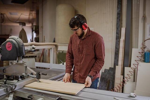Person working on a wooden board with a table saw in a woodworking workshop.