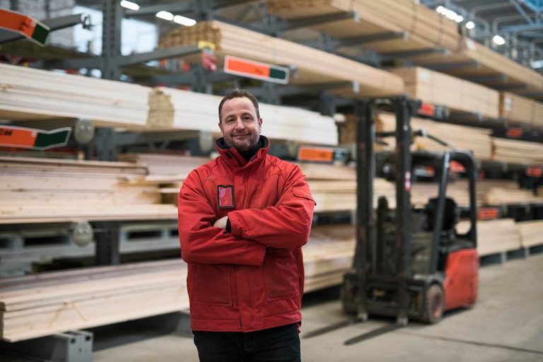 Person standing in a lumber warehouse, with stacks of wooden planks and a forklift nearby.