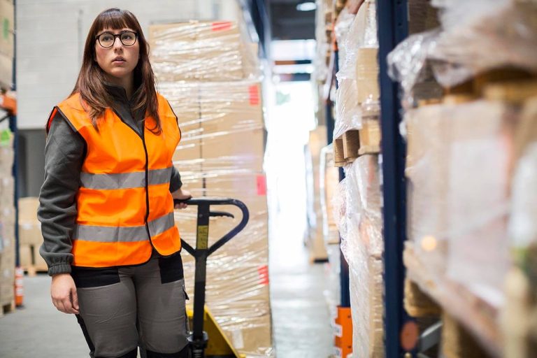 Woman in safety vest holding a pallet jack in a warehouse, surrounded by stacked boxes.