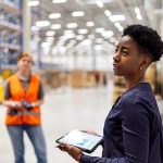 Person with tablet reviewing manufacturing processes in a warehouse, with two workers in the background.