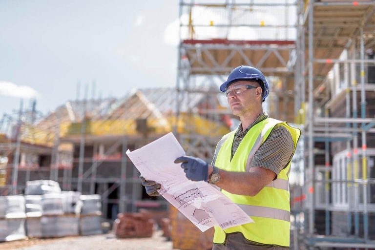 Construction worker holding a blueprint at a busy site with scaffolding and building materials.
