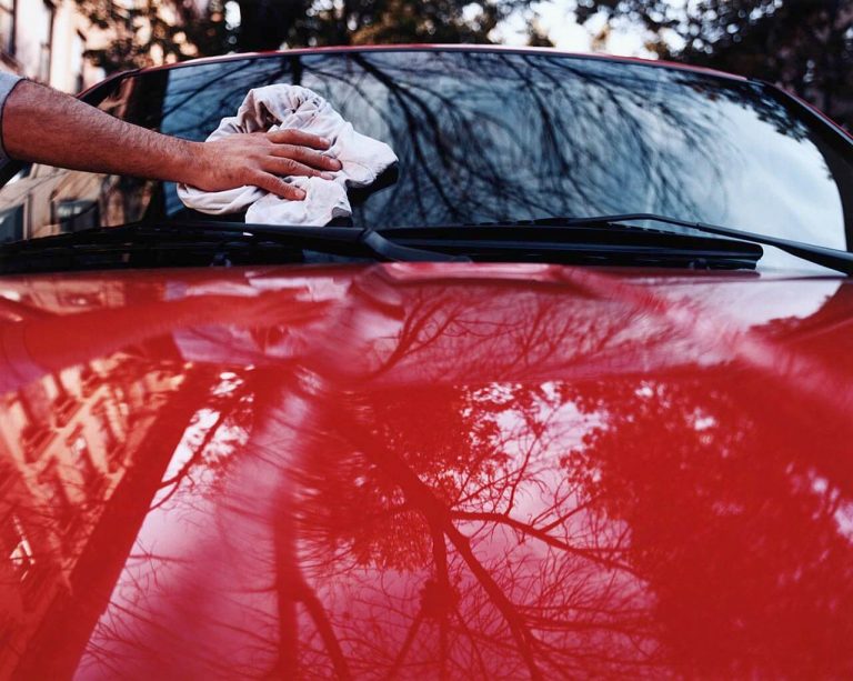 Hand cleaning a red car windshield outdoors with a cloth.. Trees and building are reflected on the car hood and windshield.